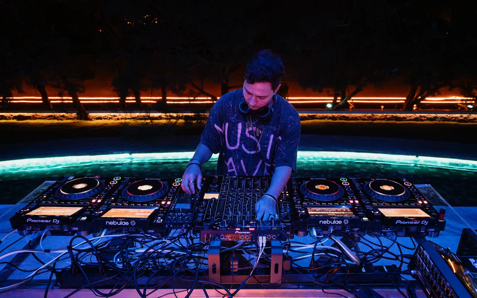 A DJ performing on a Pioneer DJ CDJ setup beside the illuminated pool at Nebulae at night